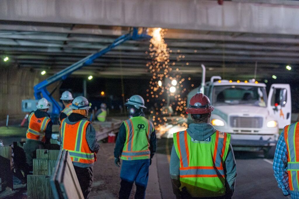 pexels photo 7976911 Nighttime road construction with workers under a bridge, sparks flying from machinery.